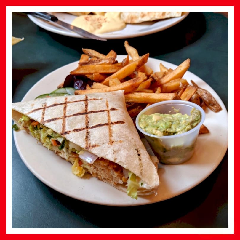 A grilled wrap filled with vegetables and chicken, served with fries and guacamole on a white plate.