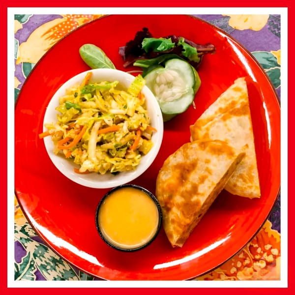 A red plate with quesadilla wedges, coleslaw, mixed greens, cucumber slices, and a dipping sauce.