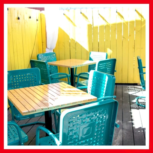 Outdoor patio with teal chairs and wooden tables against a bright yellow fence at Crazy Burger.
