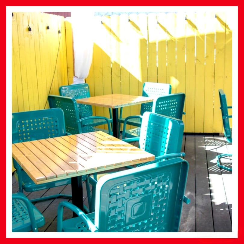 Outdoor patio with teal chairs and wooden tables against a bright yellow fence at Crazy Burger.