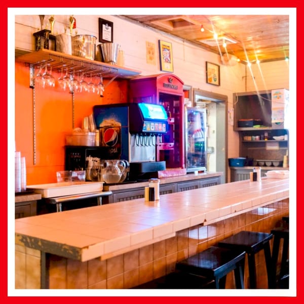 A long, tiled counter with bar stools, soda machines, and shelves with glasses at Crazy Burger.