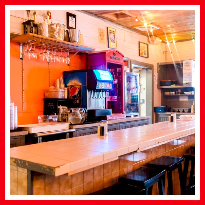 A long, tiled counter with bar stools, soda machines, and shelves with glasses at Crazy Burger.